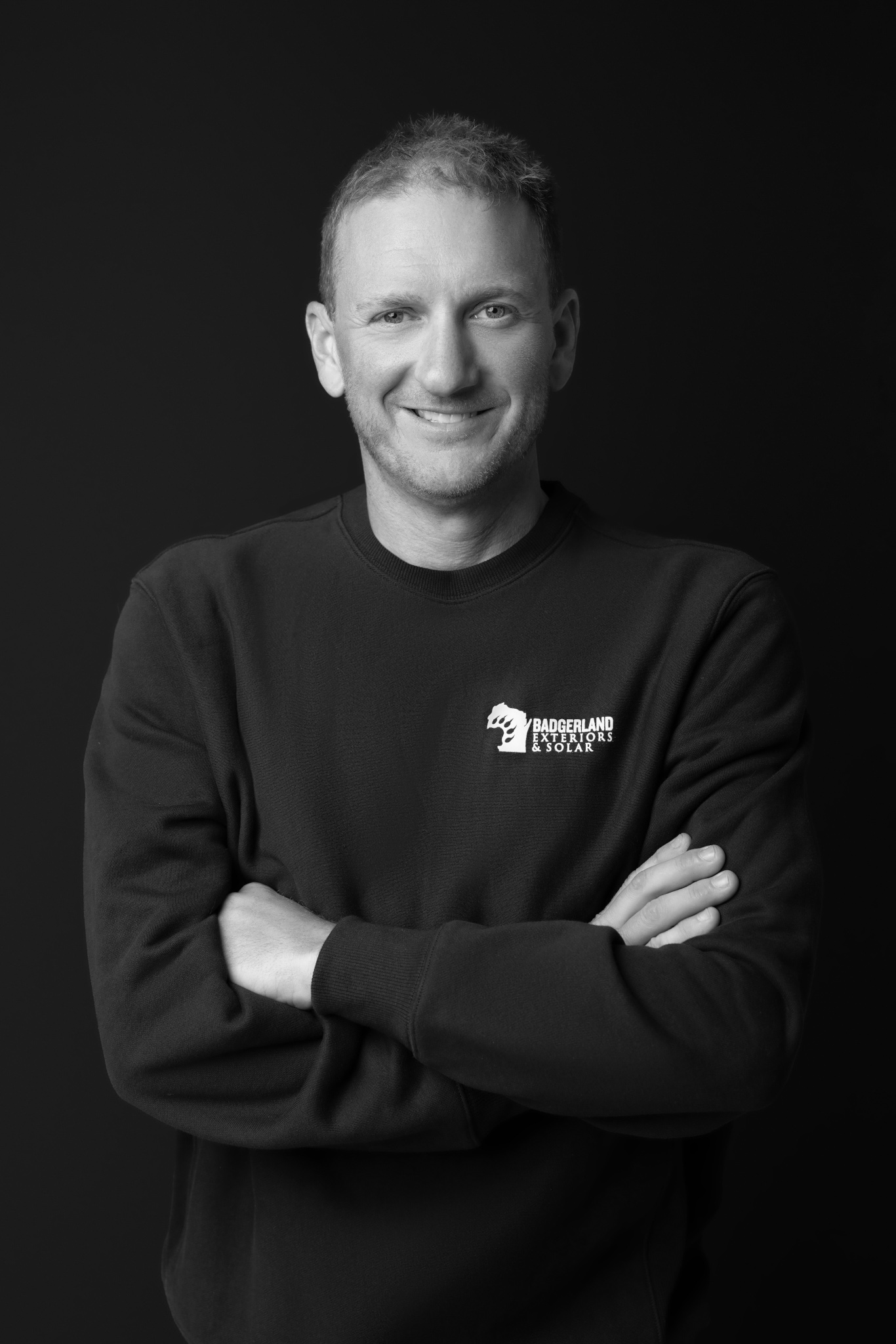 A man with short hair smiles while standing with arms crossed, wearing a dark sweatshirt with a 