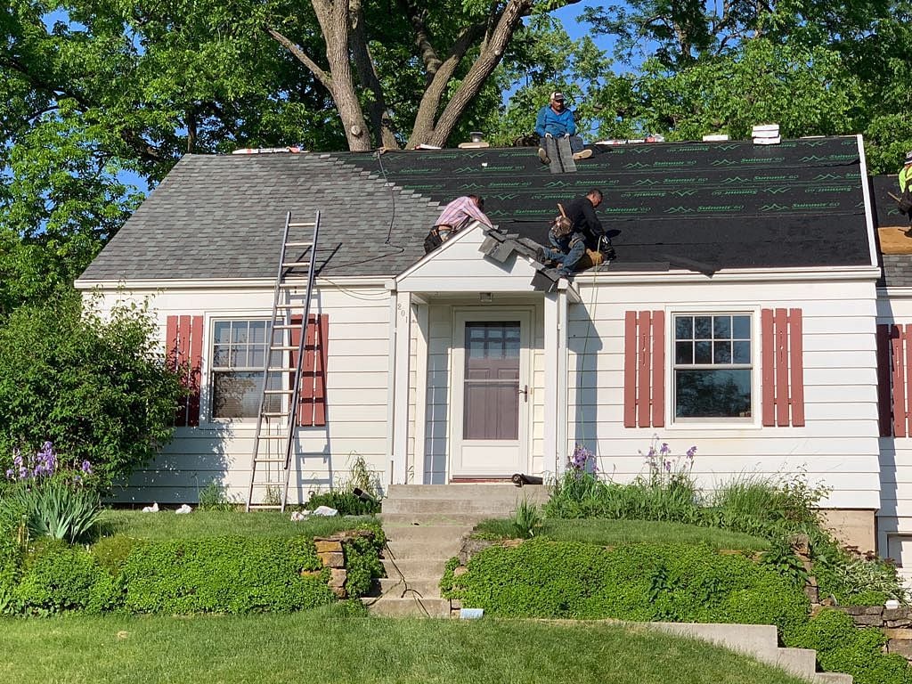 what is the most expensive part of replacing a roof in middleton wi Three workers are installing or repairing shingles on the roof of a small white house. A ladder leans against the roof, and construction materials are visible. Green bushes and flowers grow in front of the house.