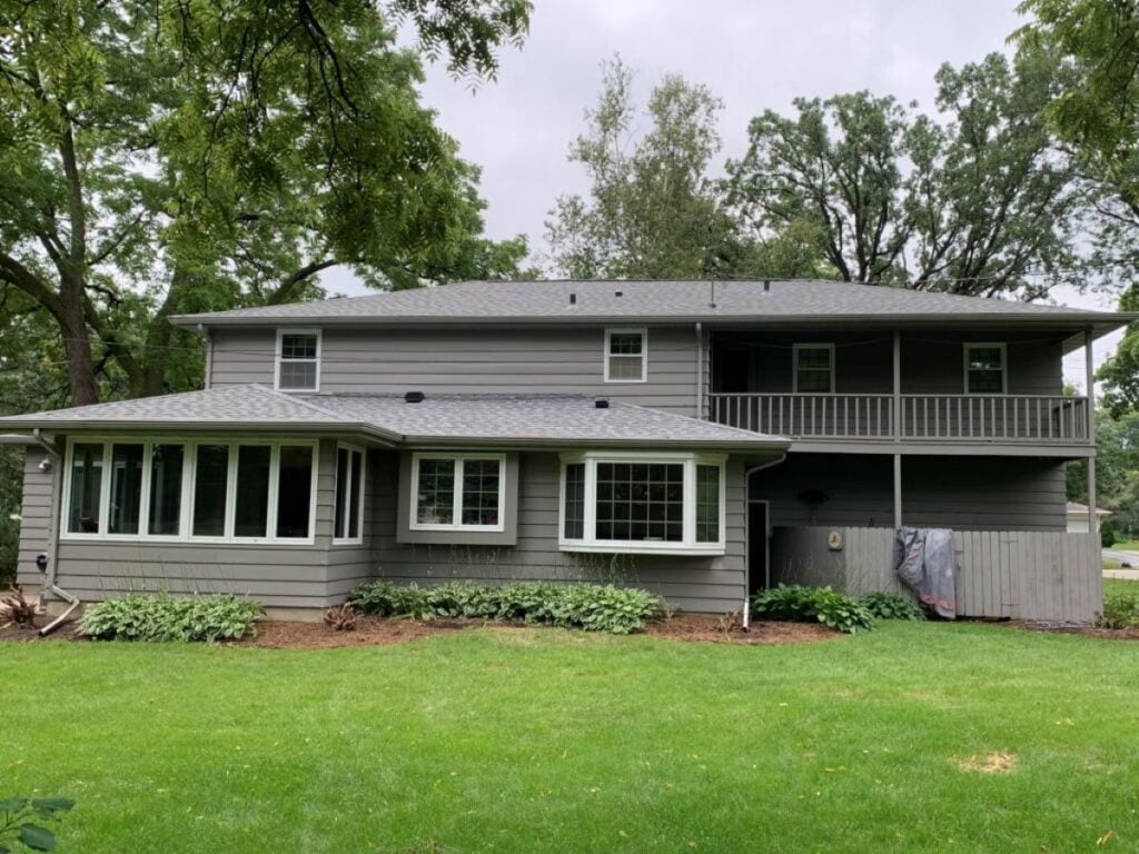 fiber cement siding installation Two-story gray house with large windows, a covered balcony, and a well-maintained lawn. Shrubs are planted along the house, and tall trees surround the area, creating a peaceful suburban setting.