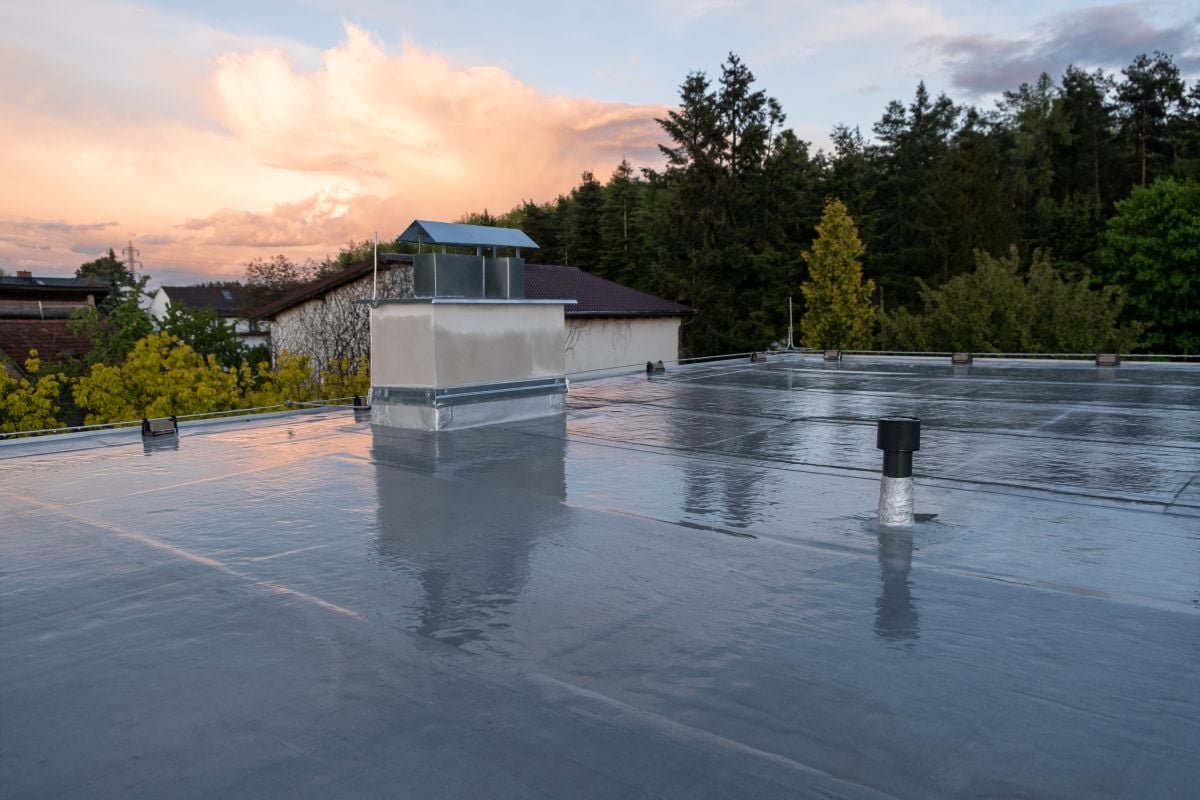 flat roof replacement A flat roof covered with a shiny, reflective waterproof membrane; several vents and a chimney are visible, with trees and a house in the background at sunset.