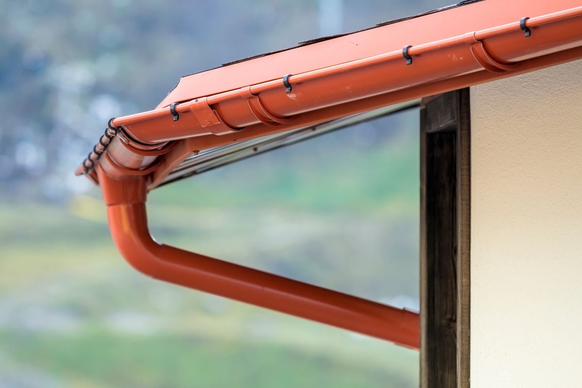 seamless gutter installation Close-up of a red rain gutter and downspout attached to the edge of a building's roof, with a blurred green and gray background.