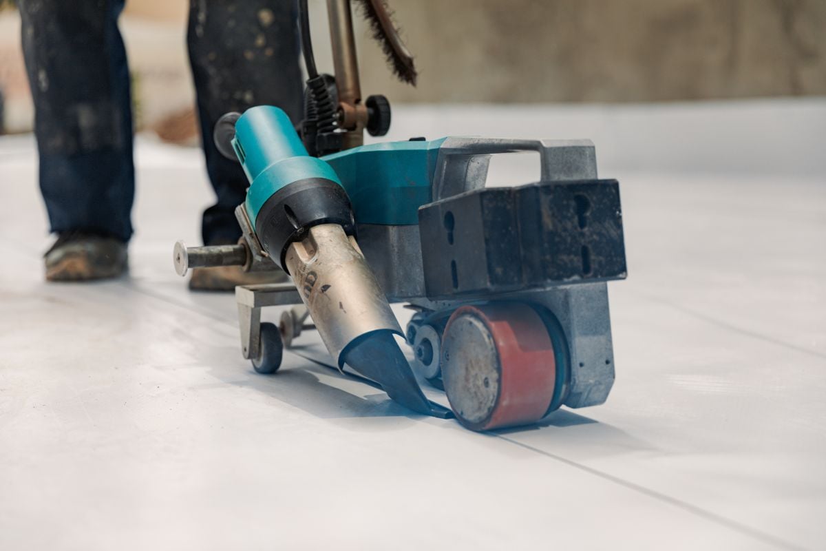 roofing companies verona wi Close-up of an automatic welding machine being used to join sheets of material on a floor, with a person's legs and work boots visible in the background.