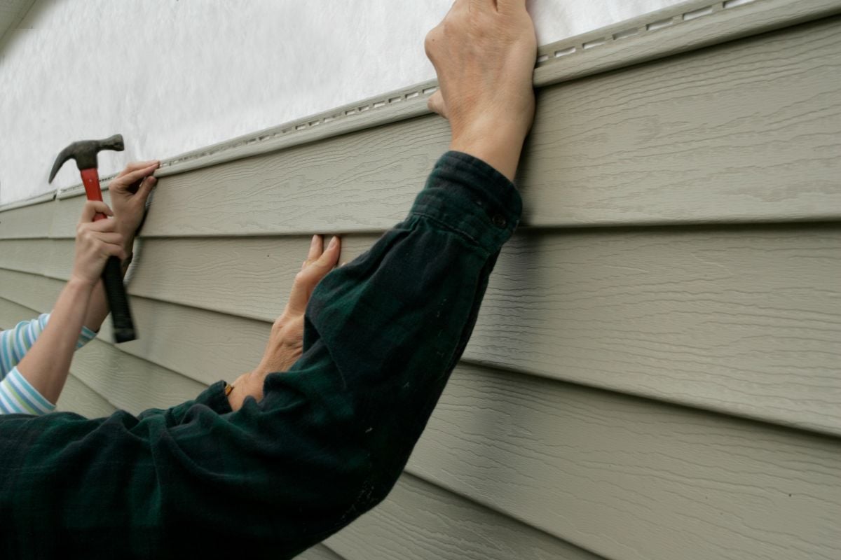 siding repair services Two people are installing light gray vinyl siding on a house; one person holds the siding in place while the other uses a hammer to secure it with nails.