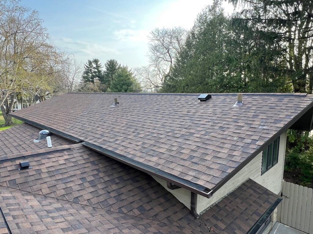 A brown shingled roof with multiple vents and chimneys, surrounded by tall trees and greenery under a partly cloudy sky.