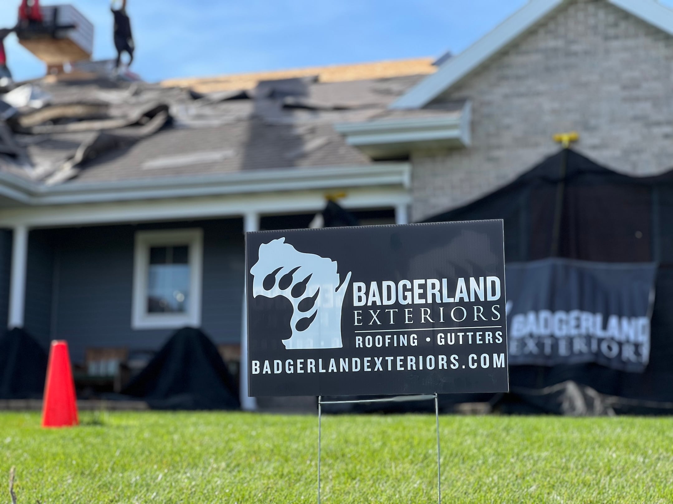 A Badgerland Exteriors sign is displayed on a lawn in front of a house undergoing roof repairs. Workers are visible on the roof, covered with tarps. The company's website is printed on the sign.