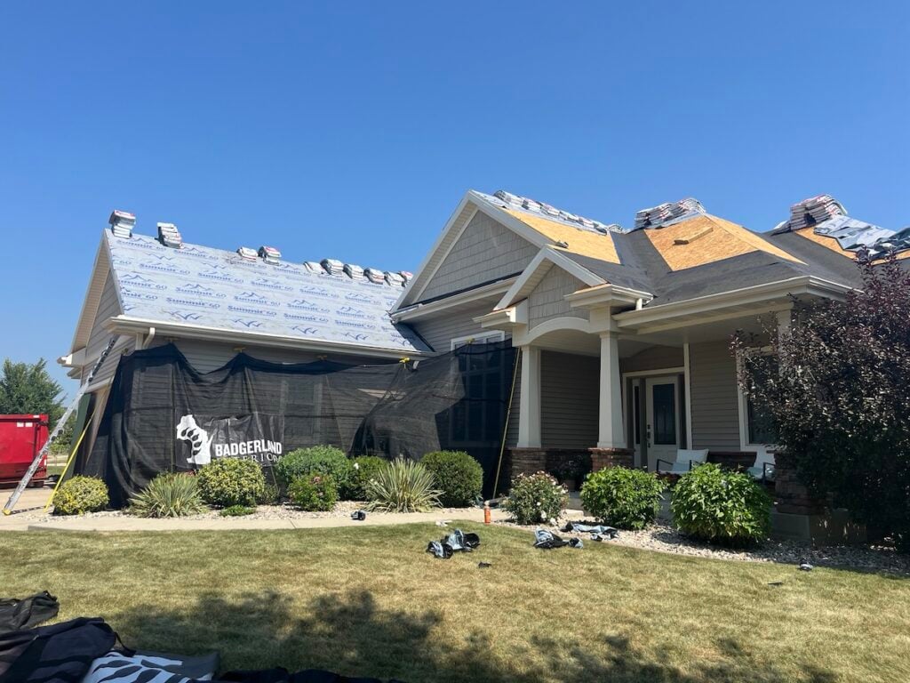 A house with roofing materials stacked on top, partial new underlayment, and some exposed sections. A black safety net and a red dumpster are visible, indicating ongoing roof construction or repair.