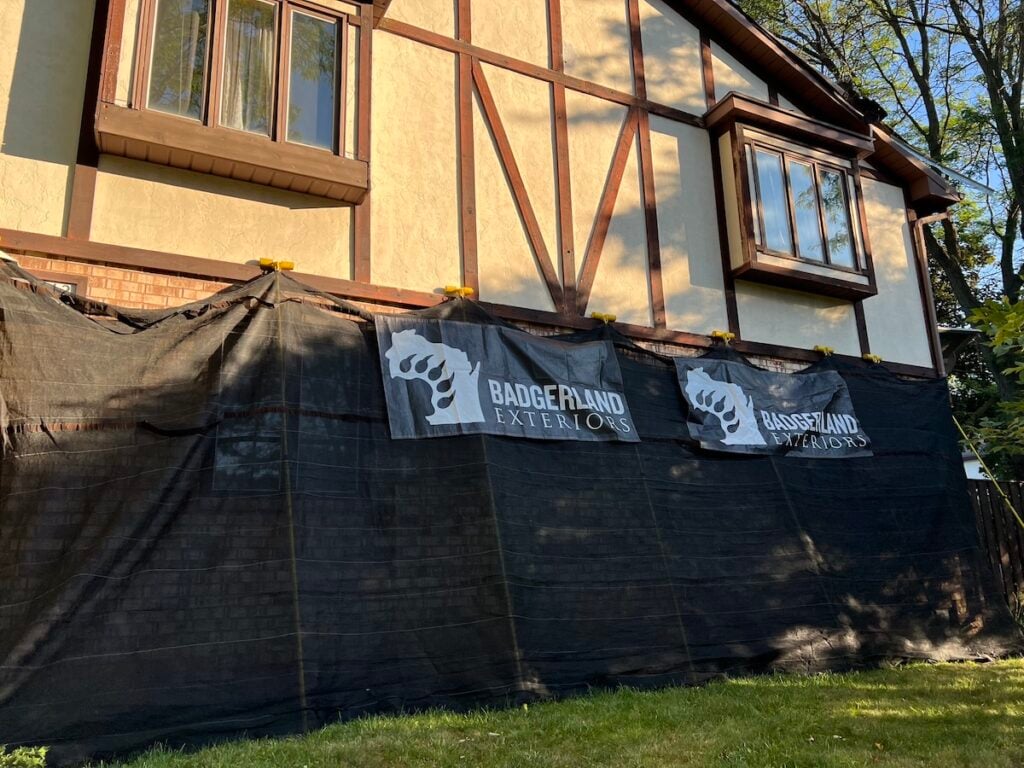 A house with Tudor-style architecture is partially covered by black construction fencing. Two "Badgerland Exteriors" banners hang on the fence. Sunlight and tree shadows are visible on the building’s facade.