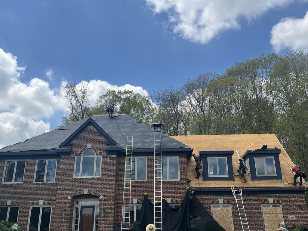 Four workers are installing or repairing the roof of a large brick house. Ladders are propped against the house, and some roofing materials are visible. The sky is partly cloudy with trees in the background.