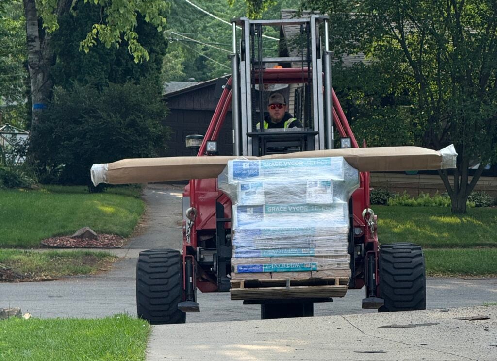 A worker drives a red forklift carrying a large pallet of construction materials, including long wrapped boards, down a residential driveway lined with green grass and trees.
