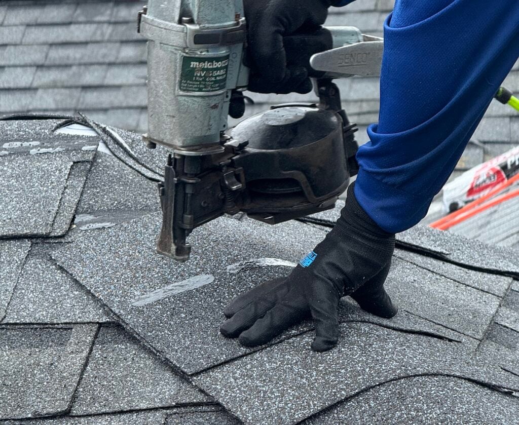 A close-up of a person in blue using a nail gun to secure gray roof shingles while wearing black gloves.
