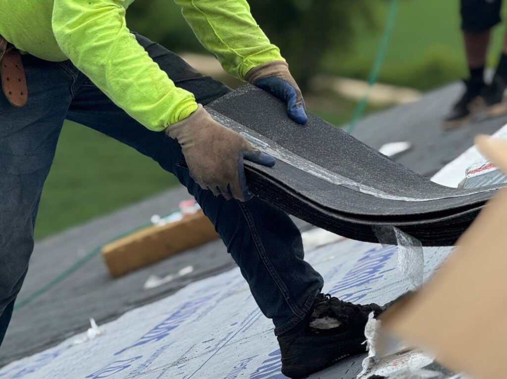 A person in a yellow shirt and gloves bends over, holding roofing shingles while working on a roof. Another person’s legs are visible in the background.