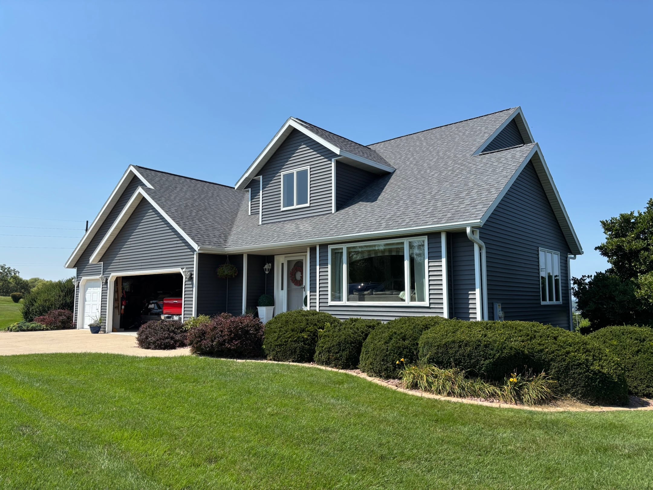 A modern gray house with white trim, a large front window, and a two-car garage sits on a manicured lawn with bushes under a clear blue sky.