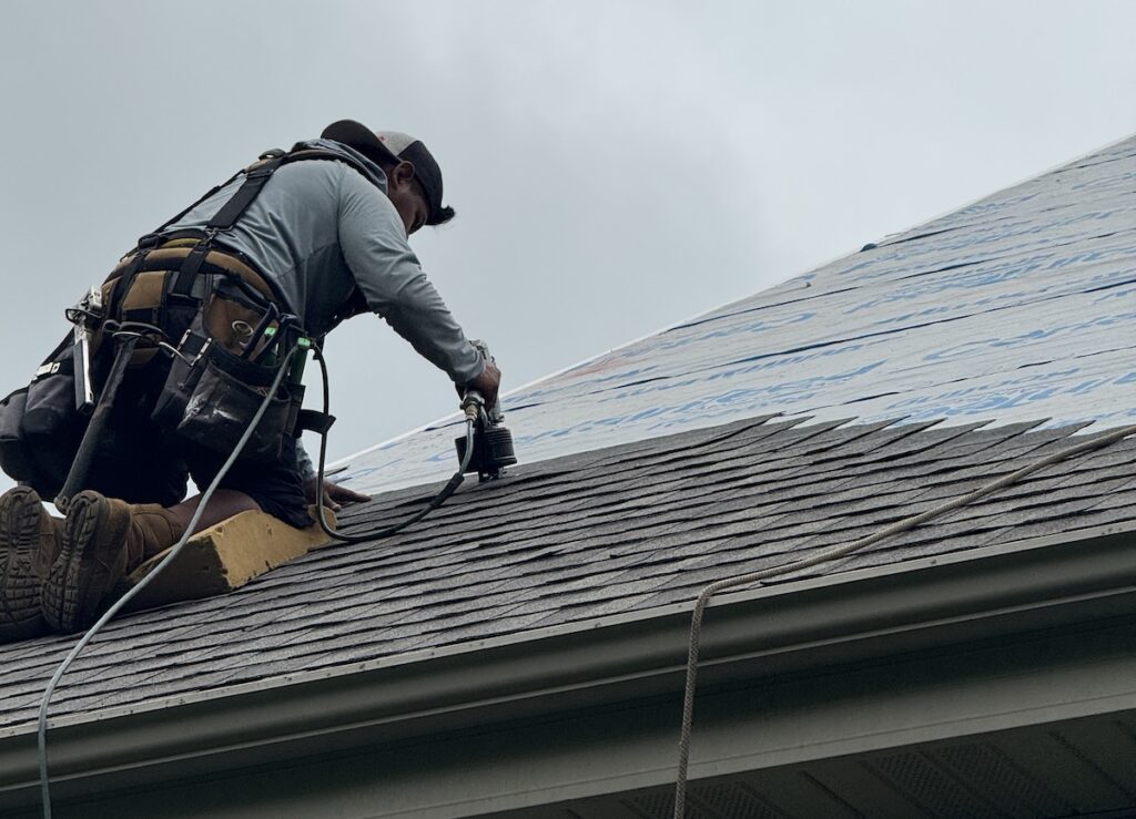 A worker wearing safety gear installs shingles on a roof using a power tool. The roof is partially covered with underlayment, and the sky is overcast.