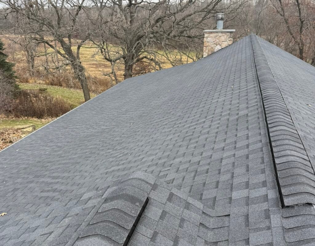 Gray asphalt shingle roof with a chimney, surrounded by bare trees and a grassy field in the background, viewed from the rooftop.