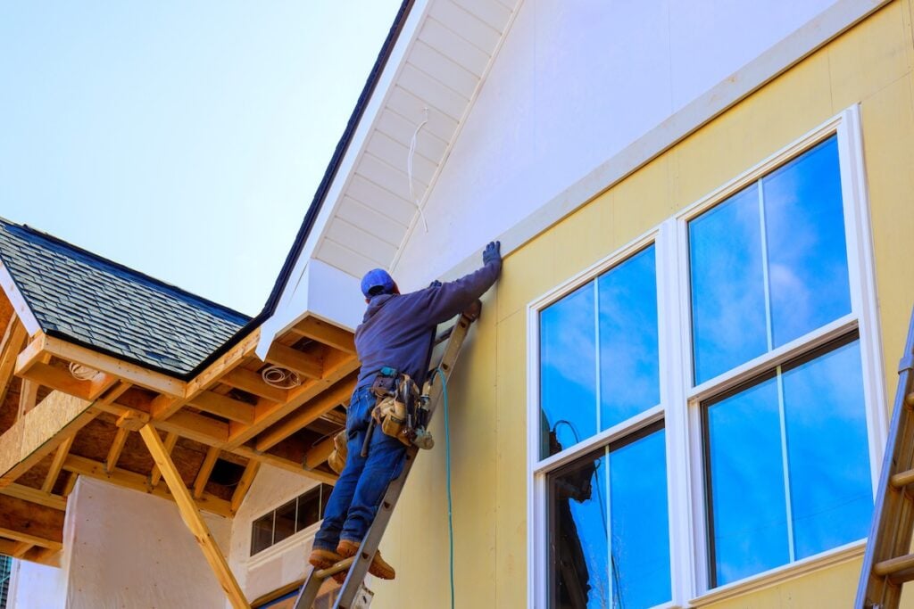 A construction worker on a ladder installs siding on a yellow house exterior near large windows, wearing a tool belt, gloves, and a hat on a clear day.