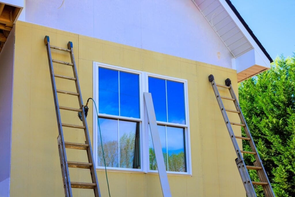 Two ladders are propped against the yellow exterior wall of a house under construction, with a double window in the center reflecting the blue sky and trees.