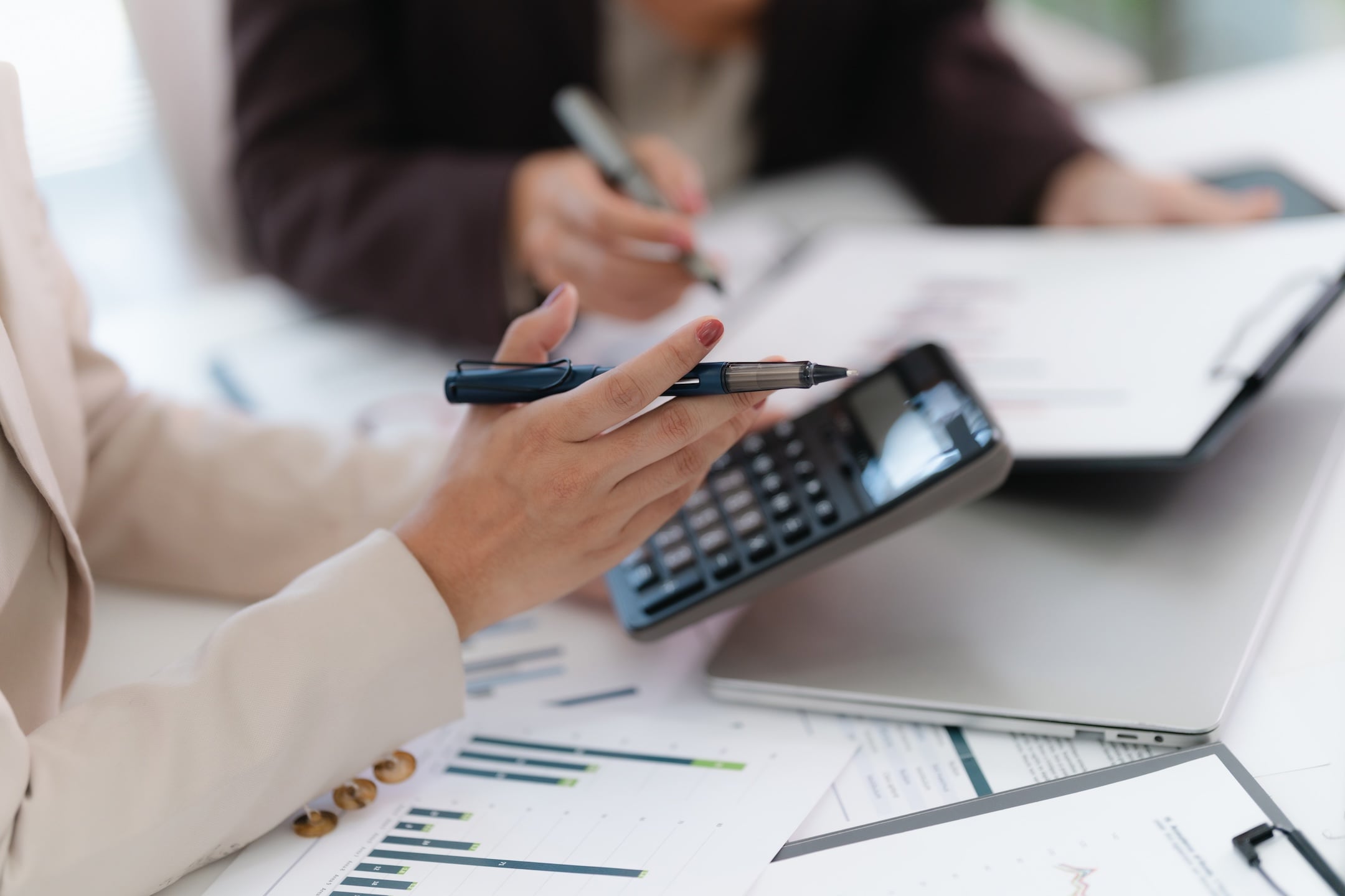 Two people discussing financial charts and graphs, one holding a pen and calculator, with papers and a laptop on the desk.