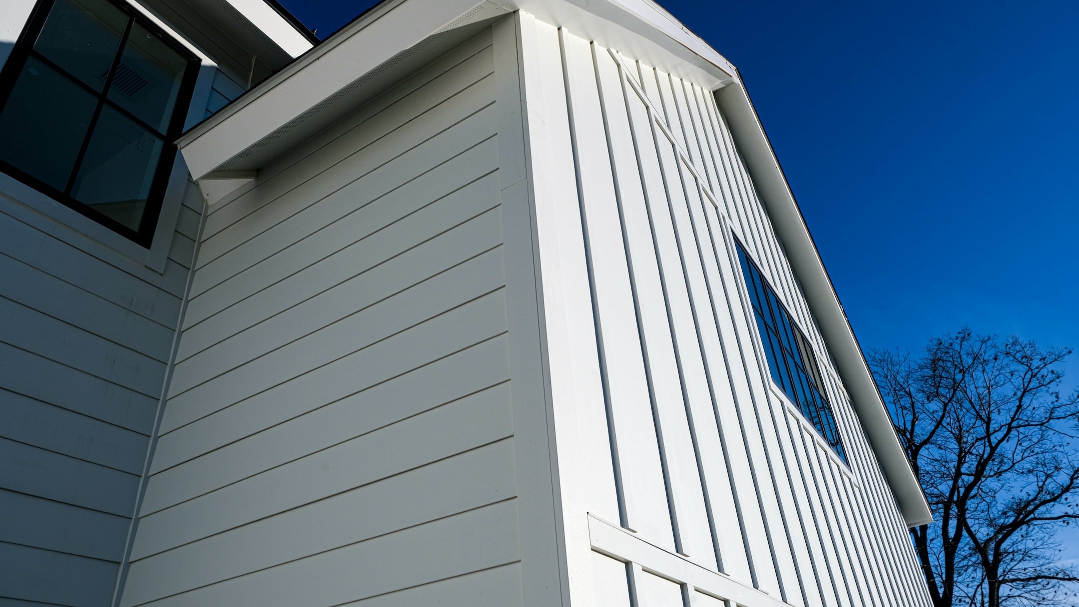 A close-up view of a modern white house exterior with vertical and horizontal siding against a clear blue sky, with a window and bare tree branches visible.