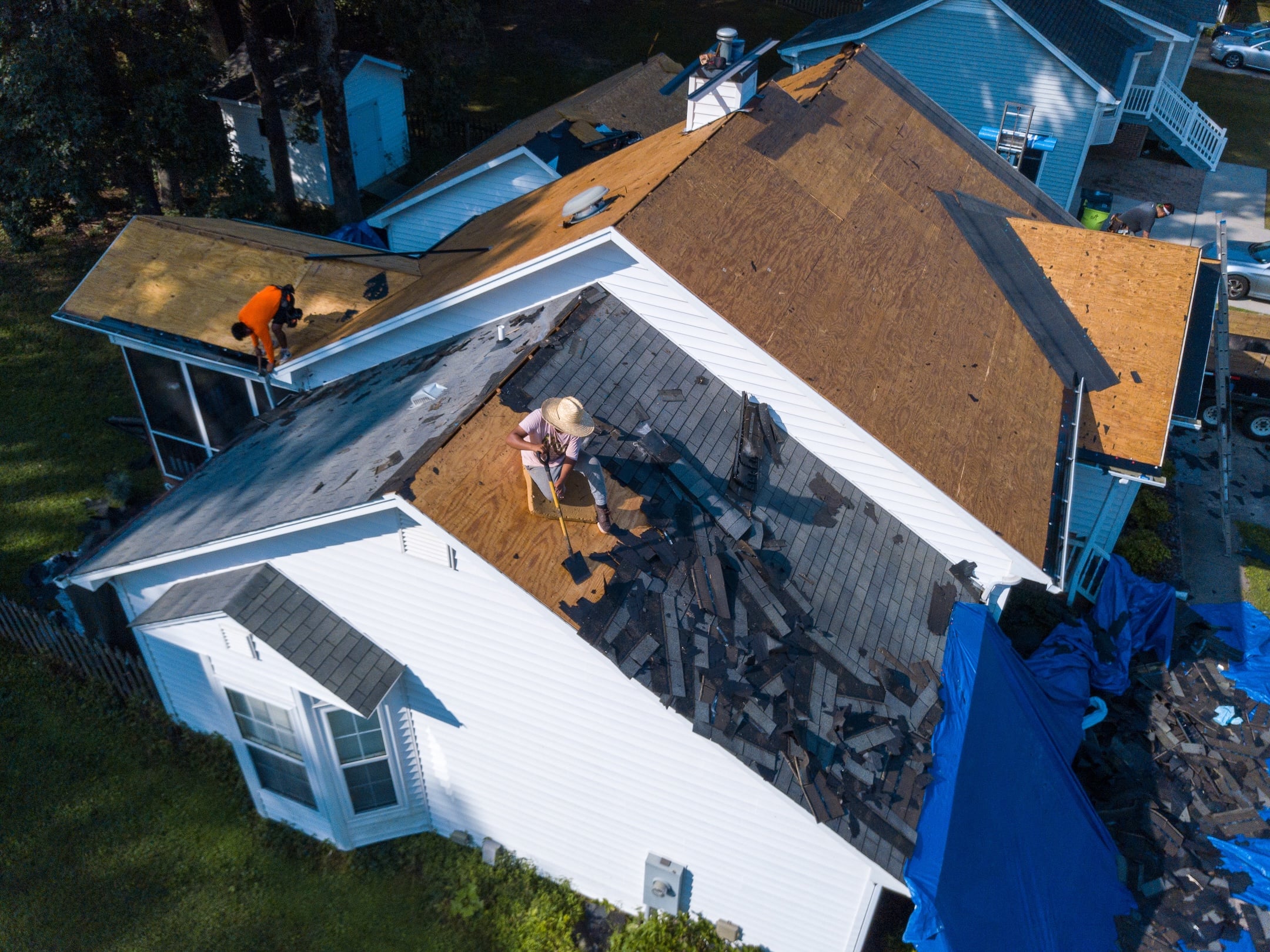 Two people work on removing old shingles from the roof of a white house. Debris and shingles are scattered on the roof and blue tarps cover sections of the ground below.