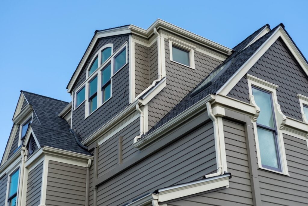 Gray and white multi-story house with various roof angles and large windows, featuring decorative fish scale siding and clear blue sky in the background.