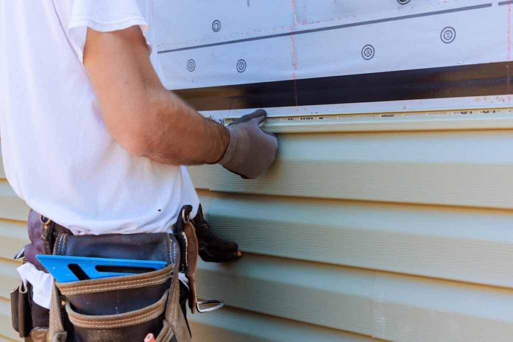 A person wearing gloves and a tool belt installs light green vinyl siding on the exterior of a building. Only their arm and torso are visible.