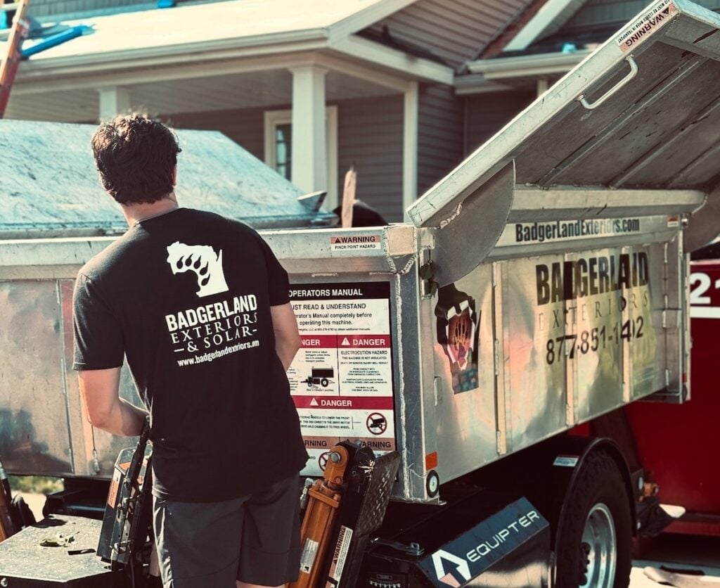 A person wearing a "Badgerland Exteriors & Solar" shirt stands next to a metal trailer with warning labels, near a house and construction materials, on a sunny day.