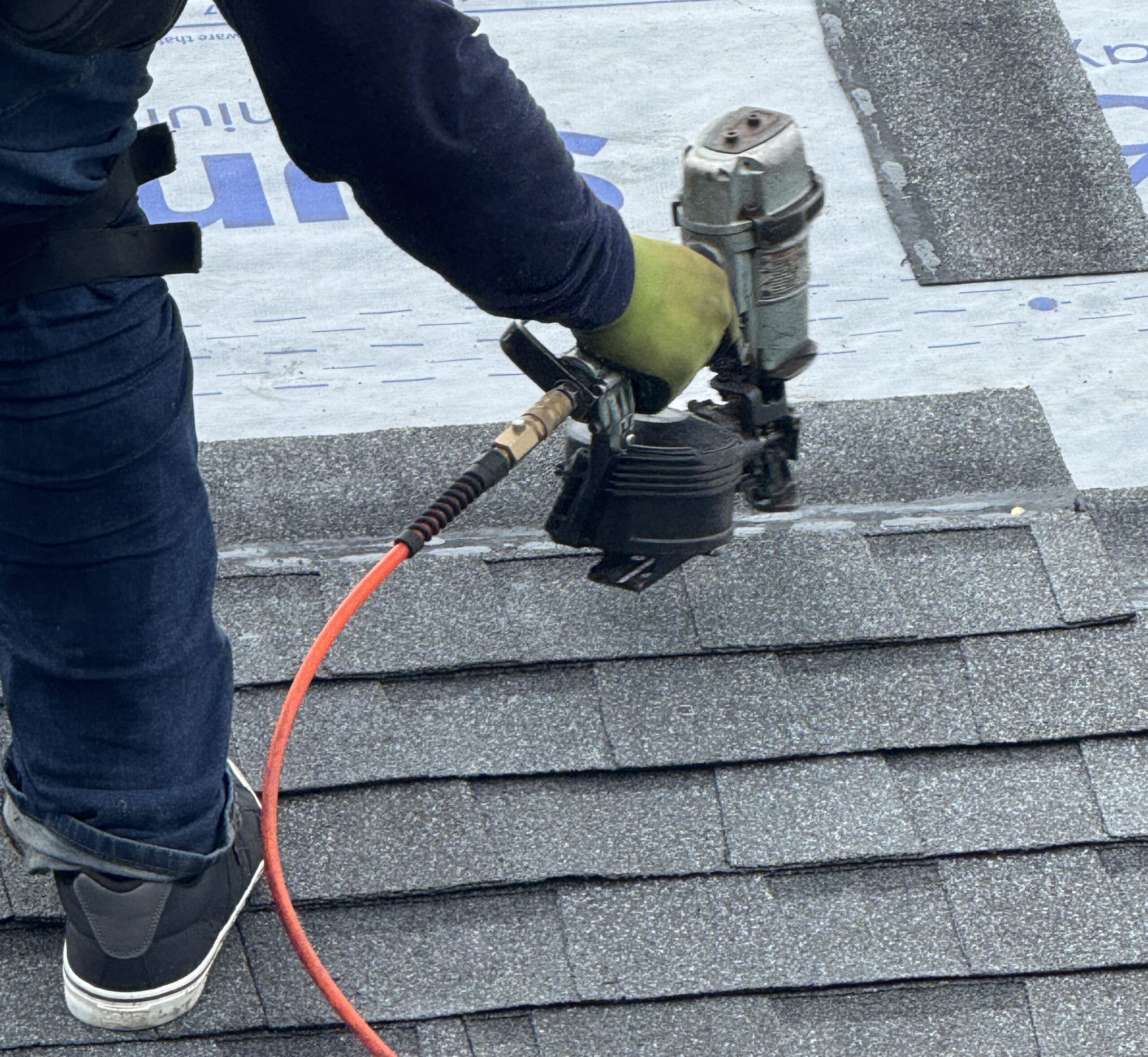 A person wearing gloves and jeans uses a pneumatic nail gun to install asphalt shingles on a roof. Roofing underlayment and shingles are visible. The worker's face is not shown.