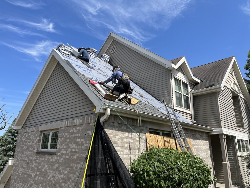 Two workers on a roof install roofing materials on a two-story house using ladders and safety gear under a clear blue sky.
