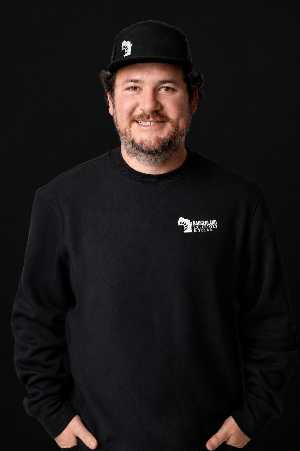 A man with a beard and mustache, wearing a black cap and black sweatshirt with a white logo, stands smiling with his hands in his pockets against a plain black background.