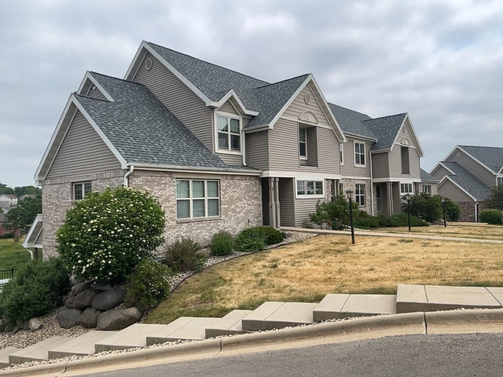 A large beige suburban house with multiple gables, gray shingles, and brick accents sits on a sloped lawn with landscaped bushes and a stone retaining wall under a cloudy sky.