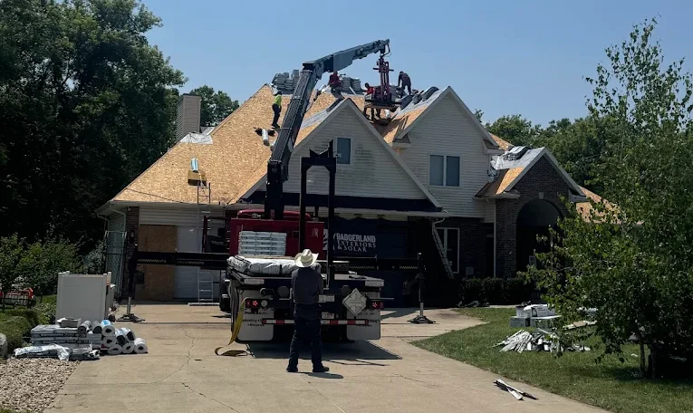Workers install new roofing on a house using a crane truck to lift materials. Roofing supplies are scattered on the driveway and lawn on a clear, sunny day.