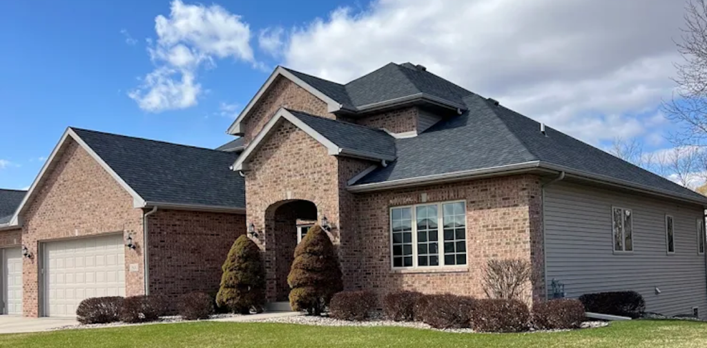 A modern brick house with a gray shingle roof, an attached two-car garage, large windows, arched entryway, and trimmed shrubs in front, under a partly cloudy blue sky.