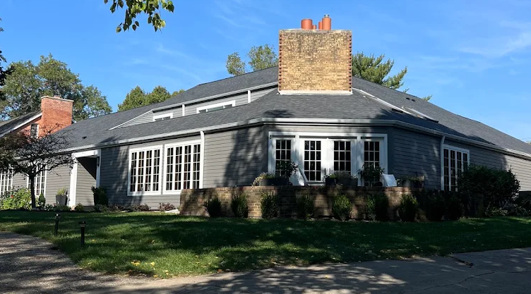 A gray, single-story house with a large brick chimney, many white-trimmed windows, and a sloped roof sits under a clear blue sky, surrounded by greenery and a paved driveway.