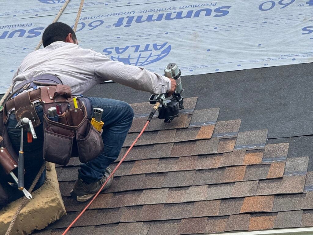 how much will it cost to fix a roof leak in waunakee wi A worker uses a nail gun to install shingles on a roof, while another person works above on underlayment. Both wear tool belts and safety gear. The image shows roofing in progress near the edge of a house.