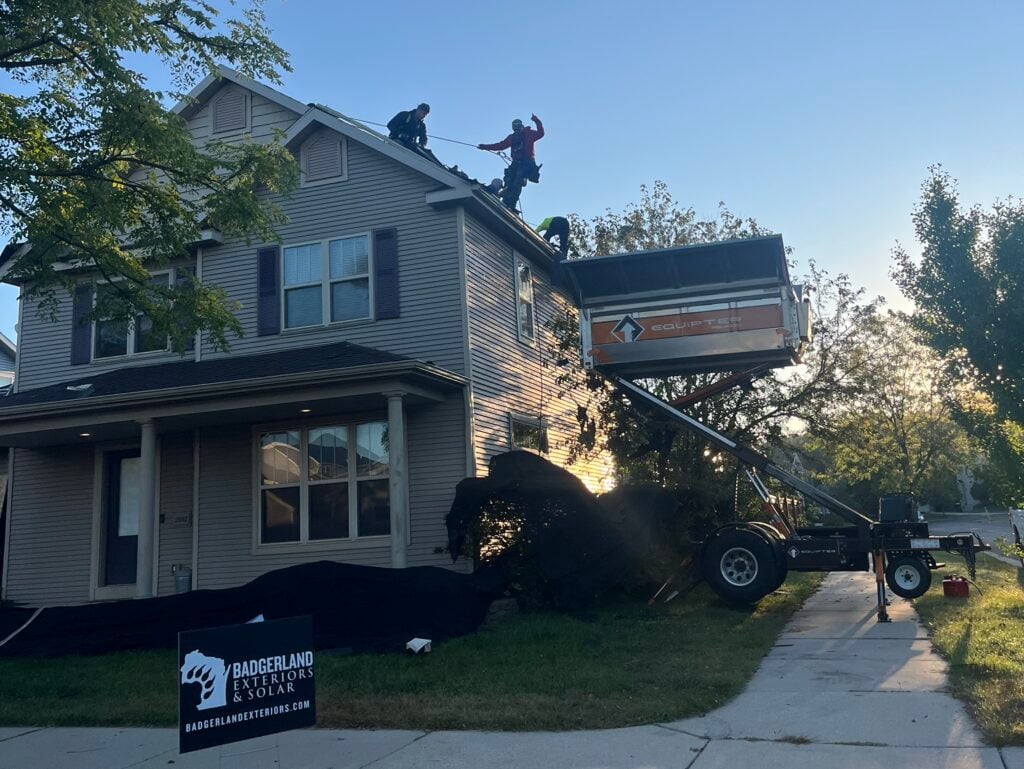 Two workers stand on a house roof, using tools. A large trailer lift is raised beside the house. Trees and a clear blue sky are in the background. A Badgerland Roofing sign is posted in the front yard.