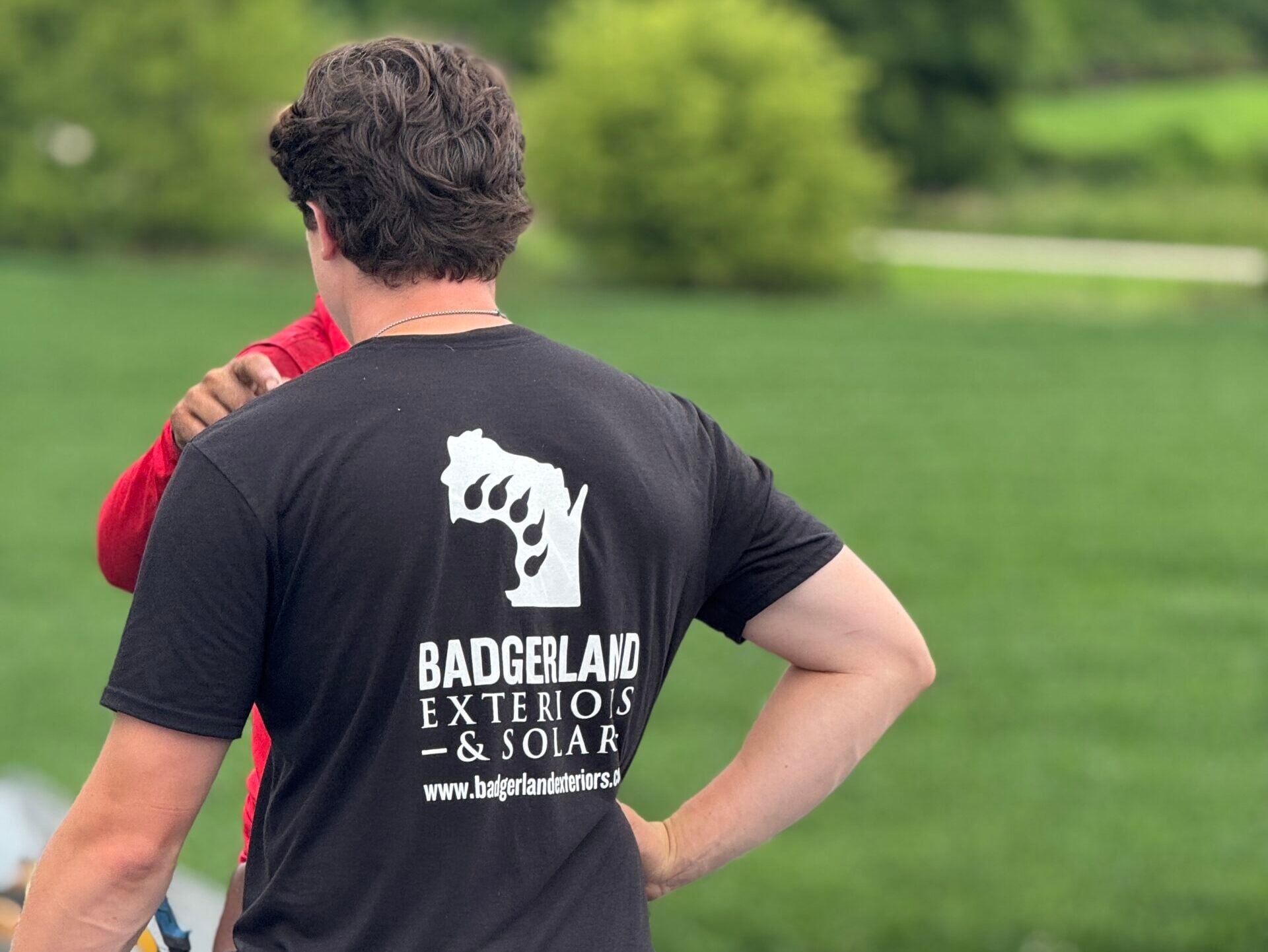 roofing companies waunakee wi workers on top of shingles roof A person in a “Badgerland Exteriors & Solar” t-shirt stands on a roof with another person, near roofing materials, with green grass and trees in the background.