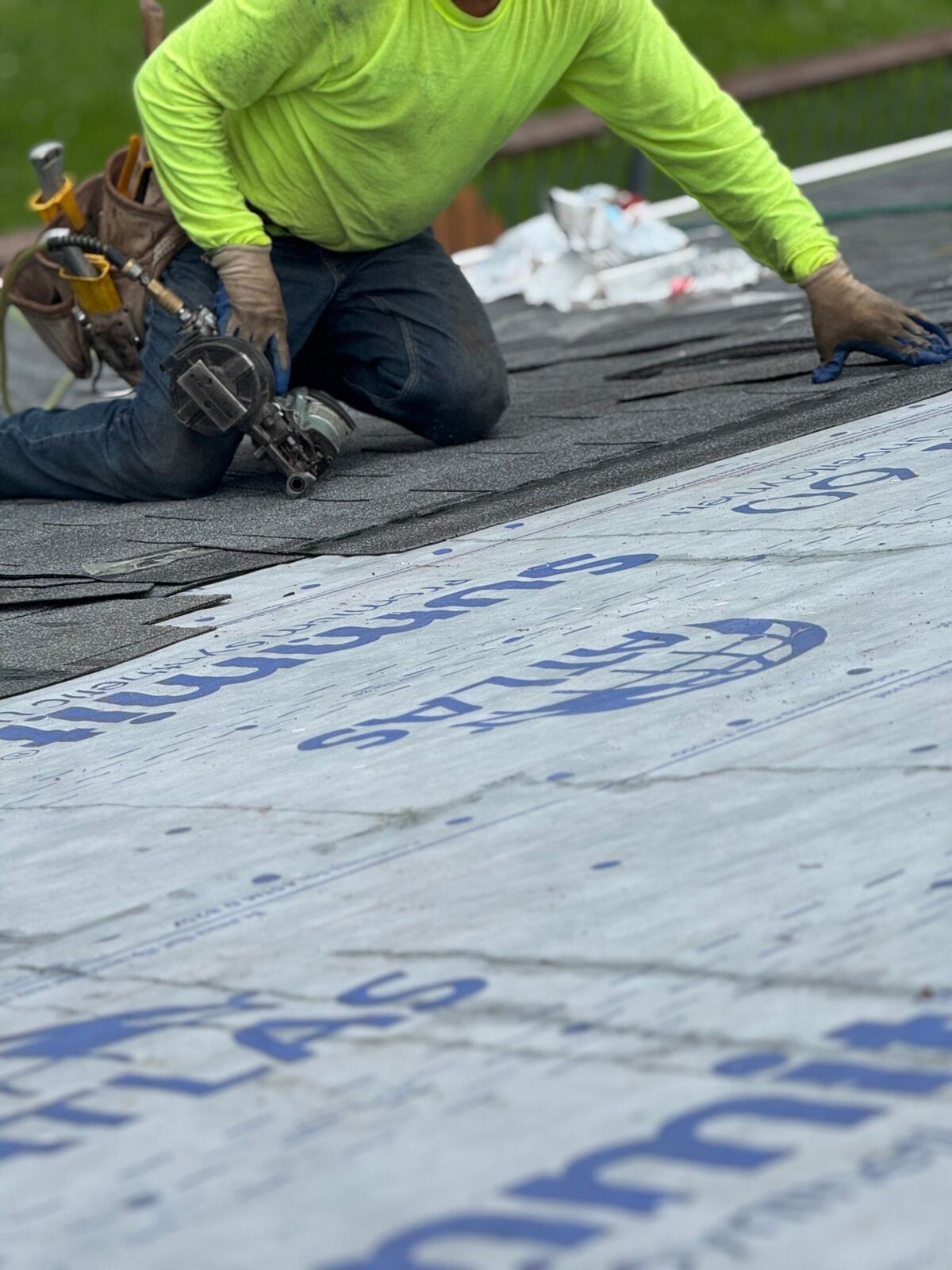 A worker in a bright yellow shirt installs roof shingles with tools nearby, kneeling on a roof covered with synthetic underlayment labeled “ATLAS.”.