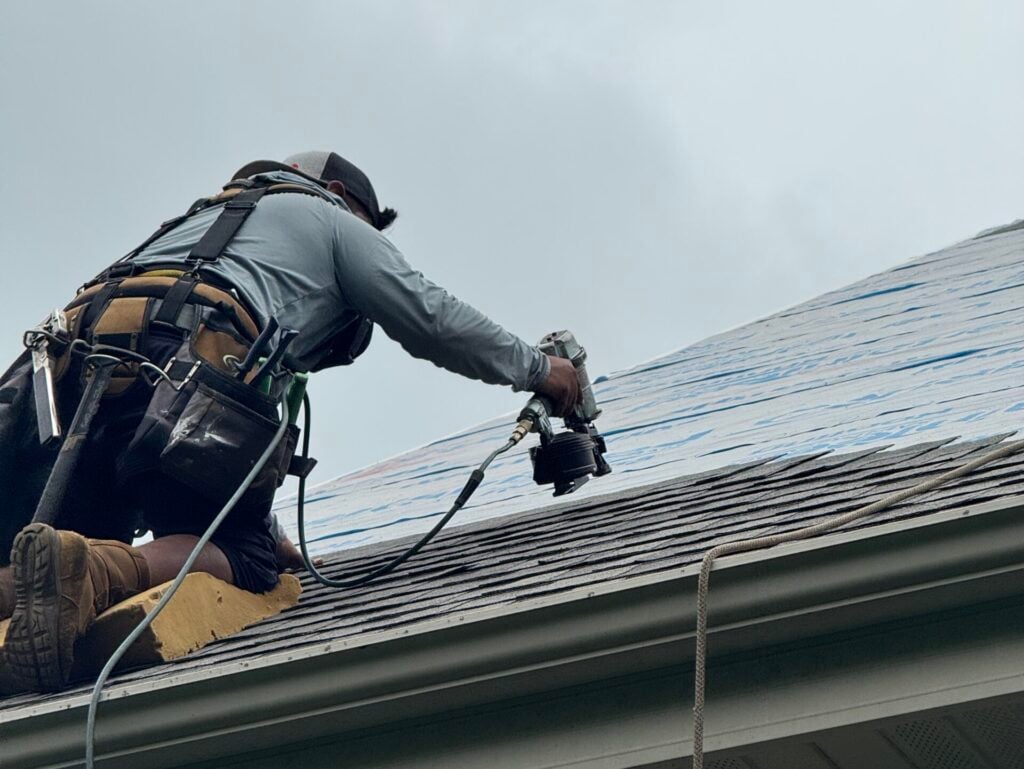 roofing companies stoughton wi A person wearing safety gear works on a house roof with tools, possibly installing shingles. The sky is overcast, and ropes are visible for safety.