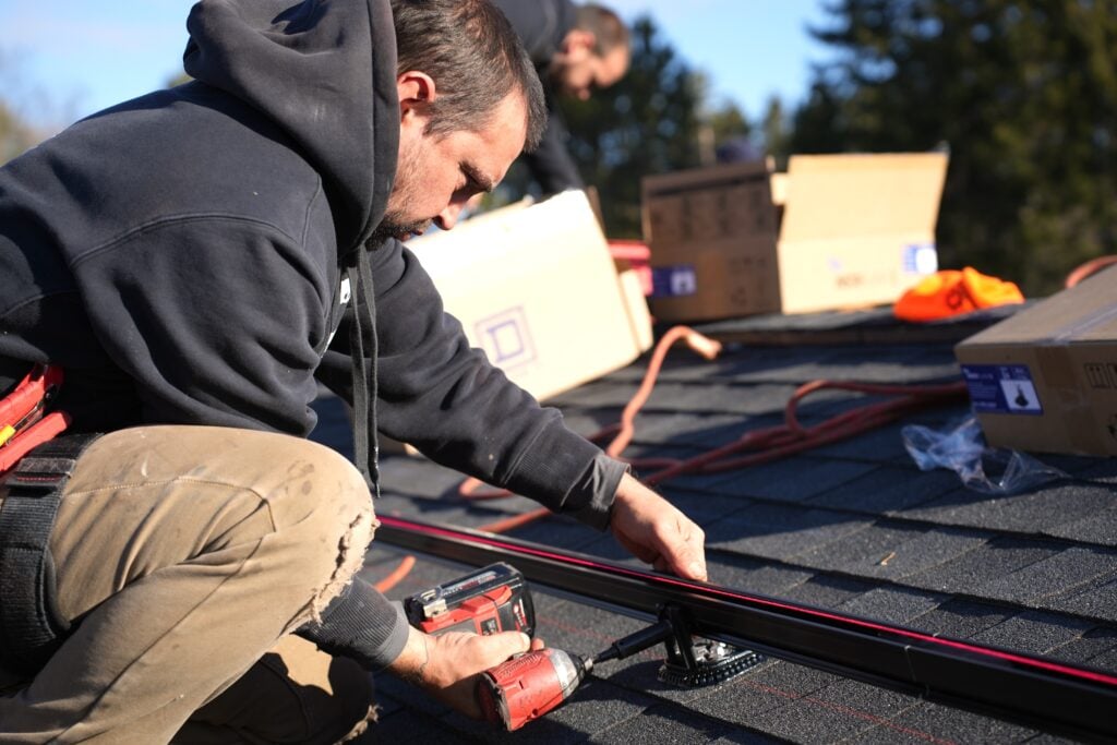 how much will it cost to fix a roof leak in waunakee wi A person in a black hoodie uses a power drill to install equipment on a shingled roof. Boxes and tools are nearby, and trees are visible in the background.
