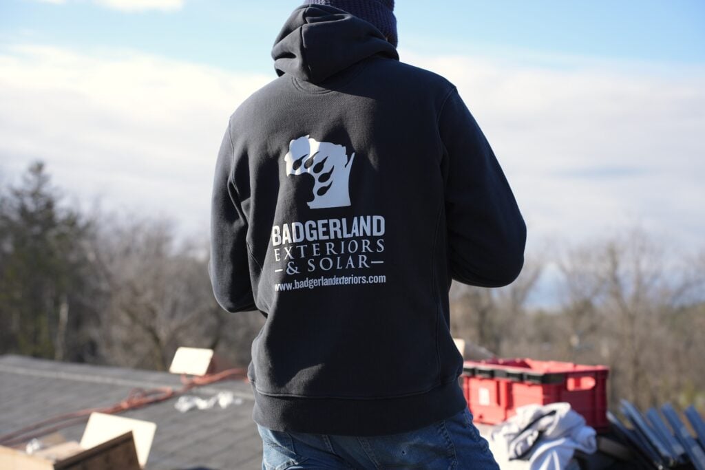 roofing companies stoughton wi Person wearing a “Badgerland Exteriors & Solar” hoodie stands outdoors with construction tools and supplies visible, facing away from the camera. Trees and cloudy sky are in the background.
