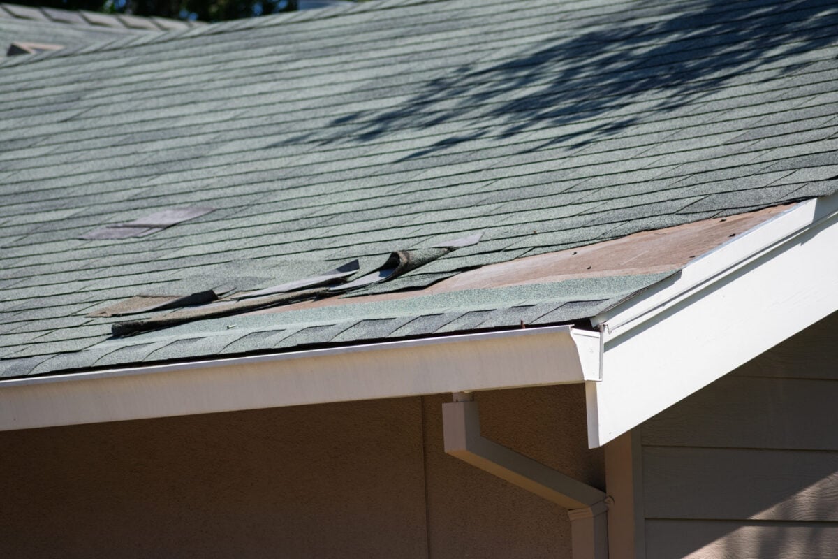 signs you need a new roof A house roof with damaged and missing shingles, exposing the underlying wood near the edge above the gutter.