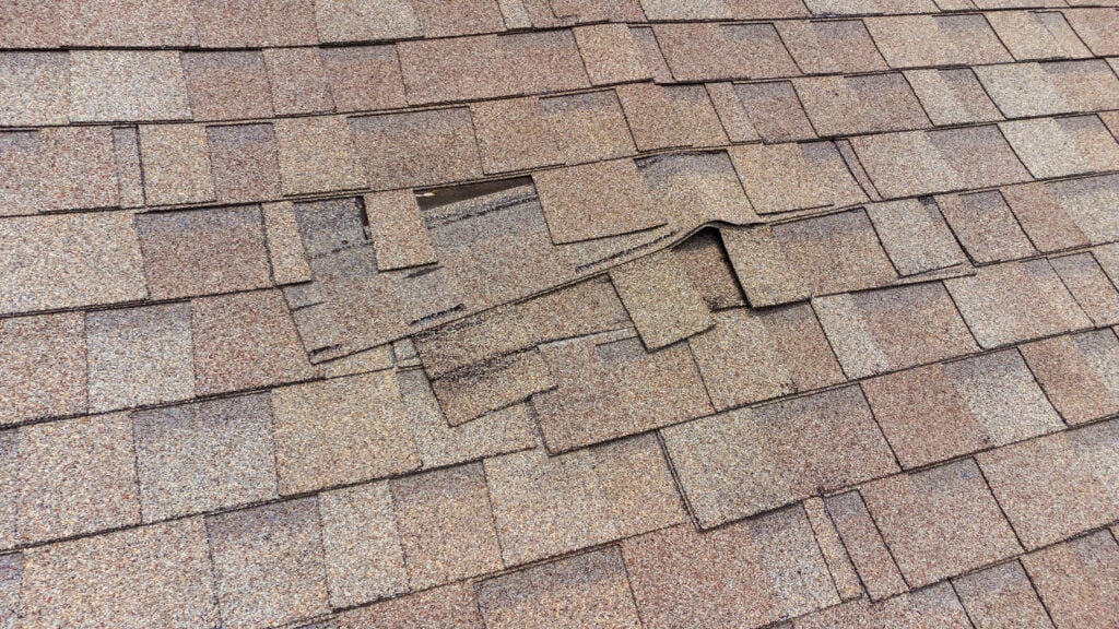 signs you need a new roof Several brown asphalt roof shingles are lifted and displaced, exposing the underlying layers, indicating damage that may need repair.