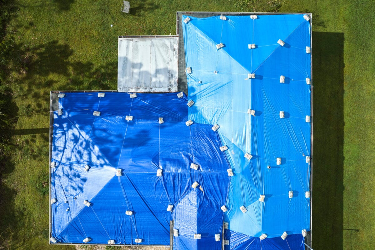 roof storm damage repair Aerial view of a house roof covered with large blue tarps secured by sandbags, surrounded by green grass and trees casting shadows.