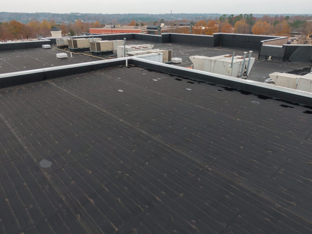 types of commercial roofing systems Flat black commercial rooftop with HVAC units and vents, surrounded by low parapet walls. Trees with autumn foliage are in the background under an overcast sky.