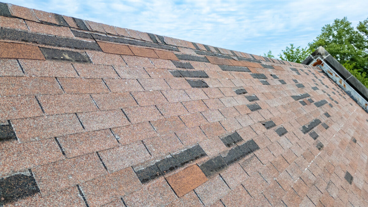 what wind speed will tear off a roof in stoughton wi Close-up of a sloped roof with brown and black asphalt shingles, some appearing worn or mismatched, under a partly cloudy blue sky with green trees visible in the background.