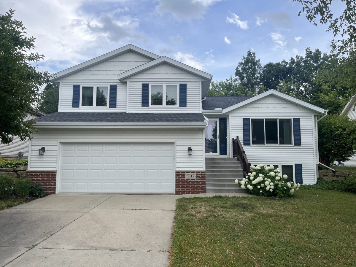 A two-story white suburban house with blue shutters, a double garage, concrete driveway, steps leading to the entrance, green lawn, and bushes with white flowers in front. Trees and blue sky are in the background.