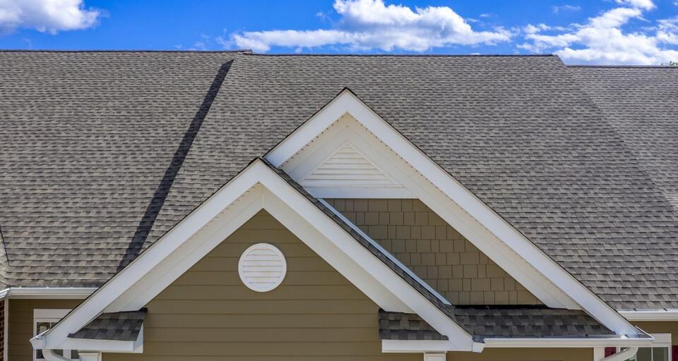 A close-up of a house roof featuring gray shingles and a gabled section with brown siding and a small round vent.