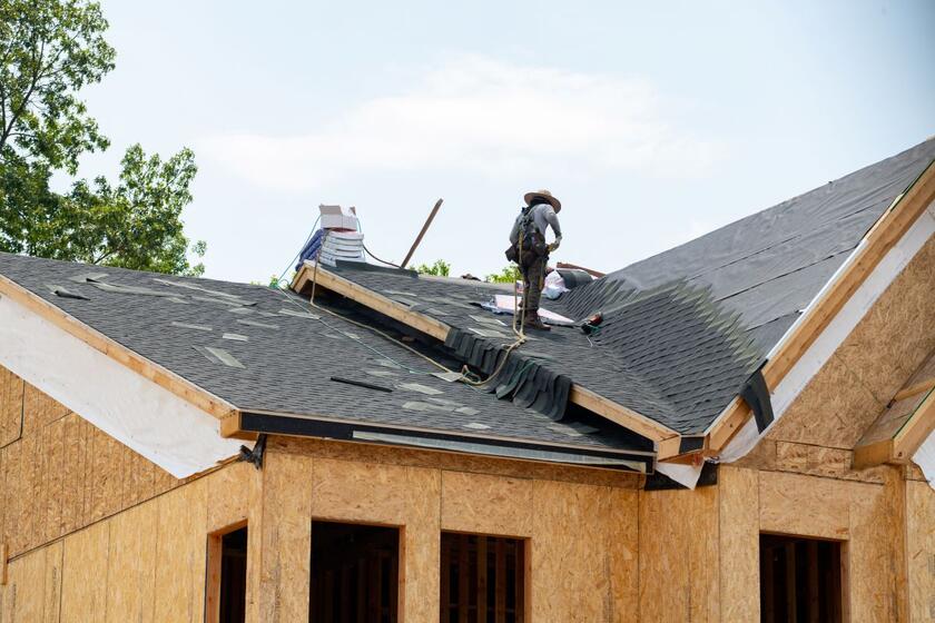 A roofer wearing a hat and safety harness works on the roof of a house under construction.